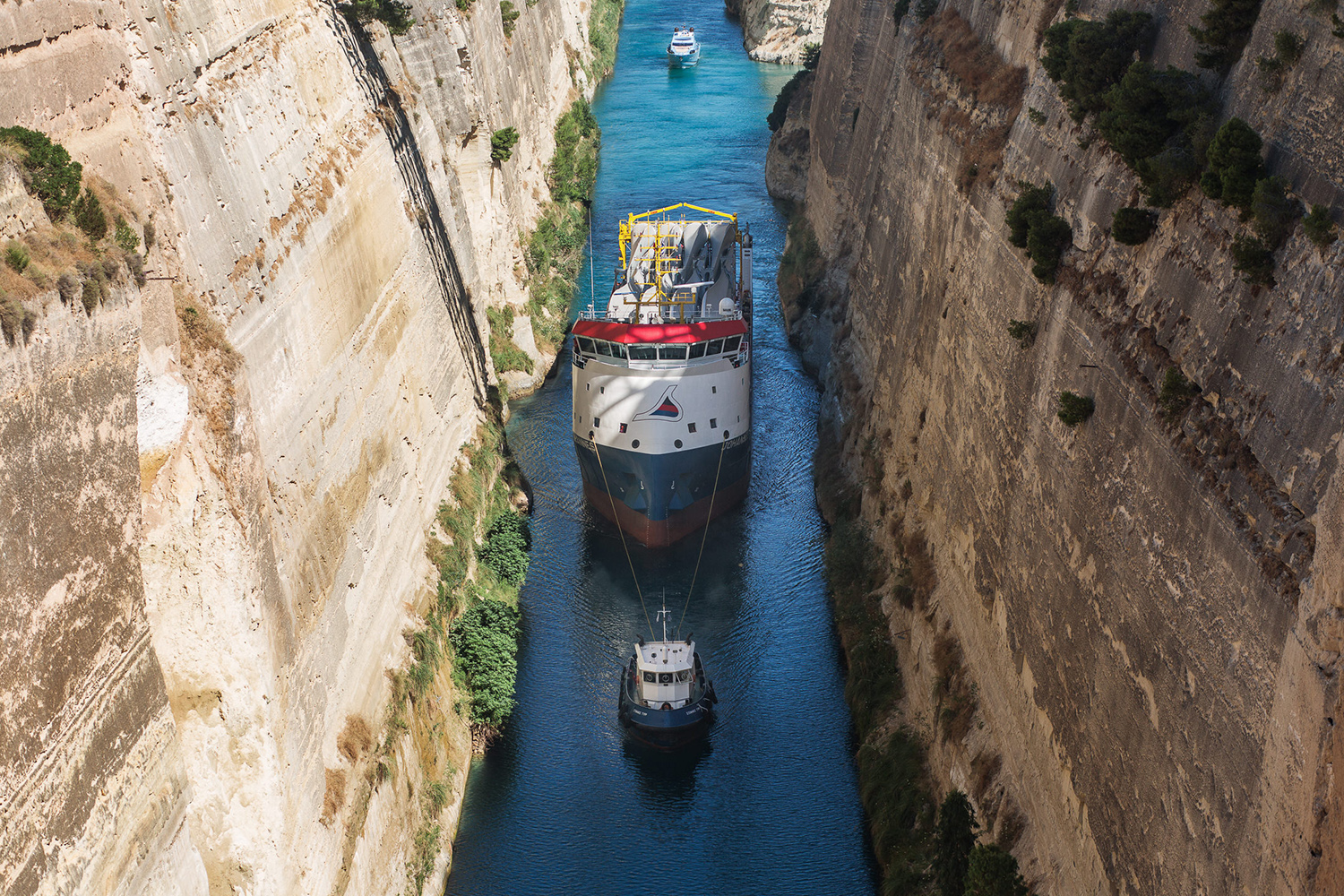 A large cargo ship being guided by a tugboat through a narrow canal with steep rocky walls, and another smaller vessel in the background.